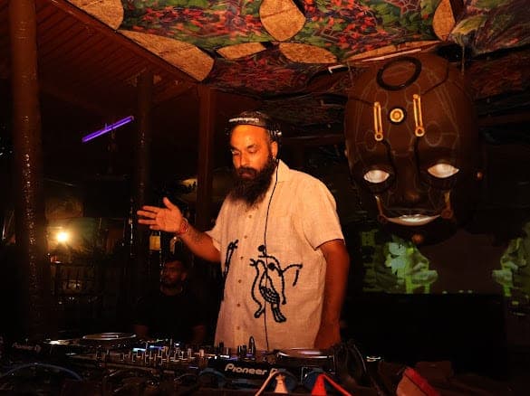 Immortal Baba behind the CDJs beneath a hand-painted canopy, gesturing towards the floor