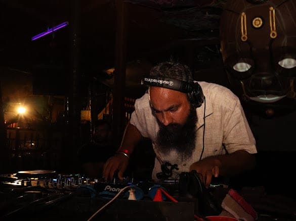 Immortal Baba bent over the mixer in near-darkness, headphones on