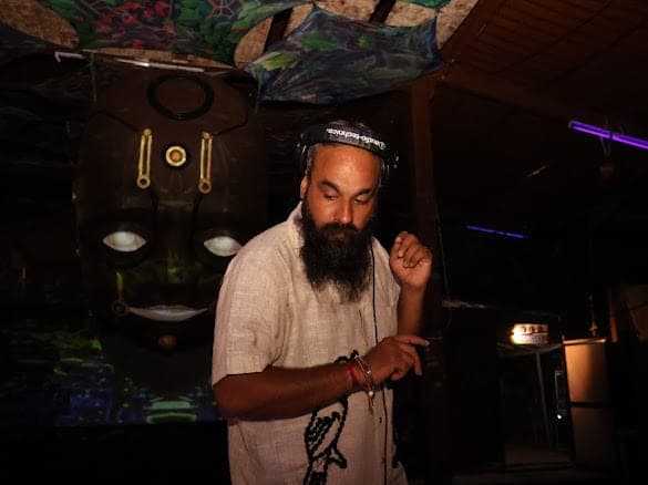 Immortal Baba at the decks beside a large carved mask with glowing eyes, a violet laser overhead